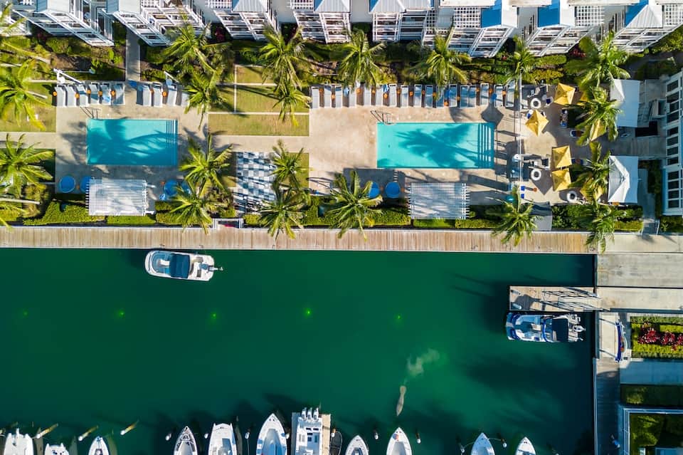 Overhead view of waterfront resort with pools, loungers, palm trees, boats, and green waterway