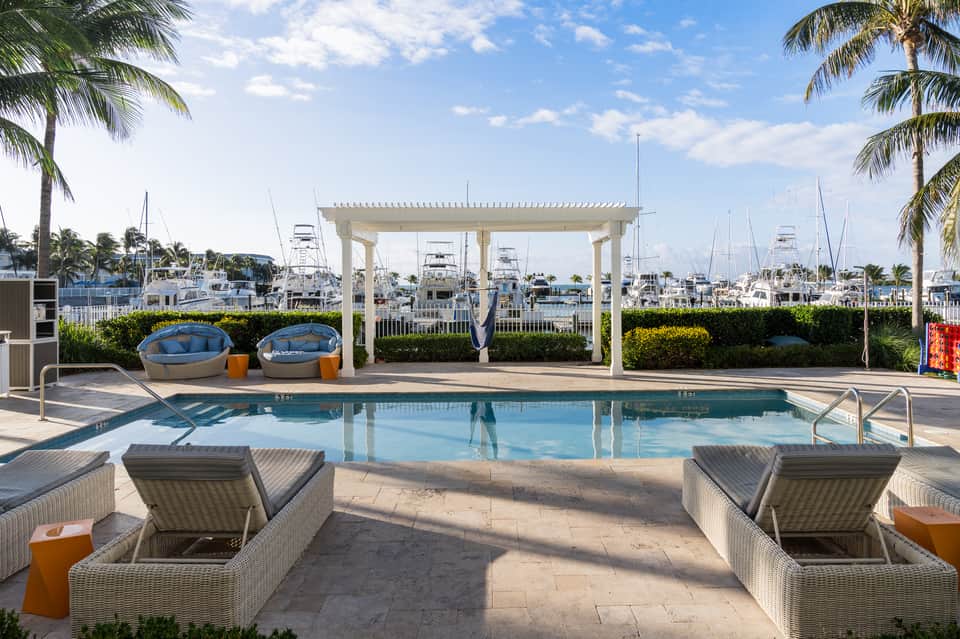 Waterfront pool deck with lounge chairs, white pergola, marina boats, and palm trees