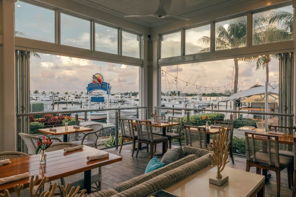 Waterfront dining area with glass walls overlooking marina and sailboats at dusk