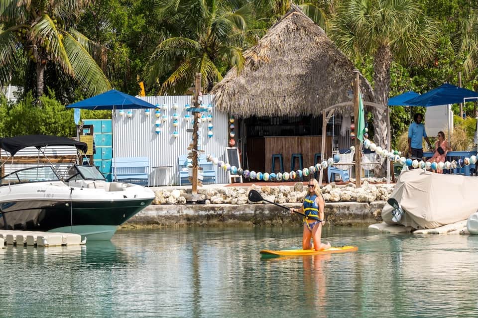 Beachfront cabana with thatched roof, white railings, blue umbrellas, and water sports activities in turquoise lagoon