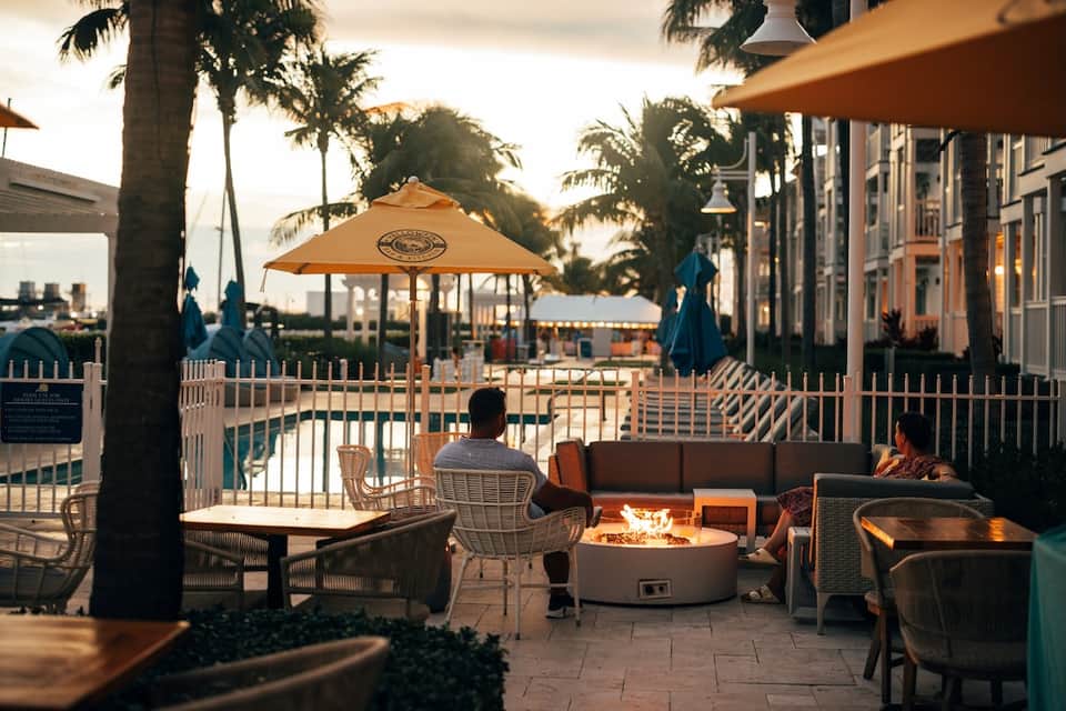 Poolside fire pit lounge at sunset with blue umbrellas, palm trees, bar, and waterfront in background