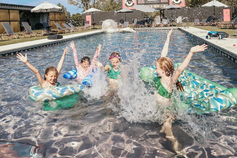 Children playing and splashing in resort swimming pool with floats, loungers, and stage visible