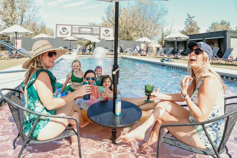 Family and friends enjoying poolside gathering with drinks at table near resort pool and buildings