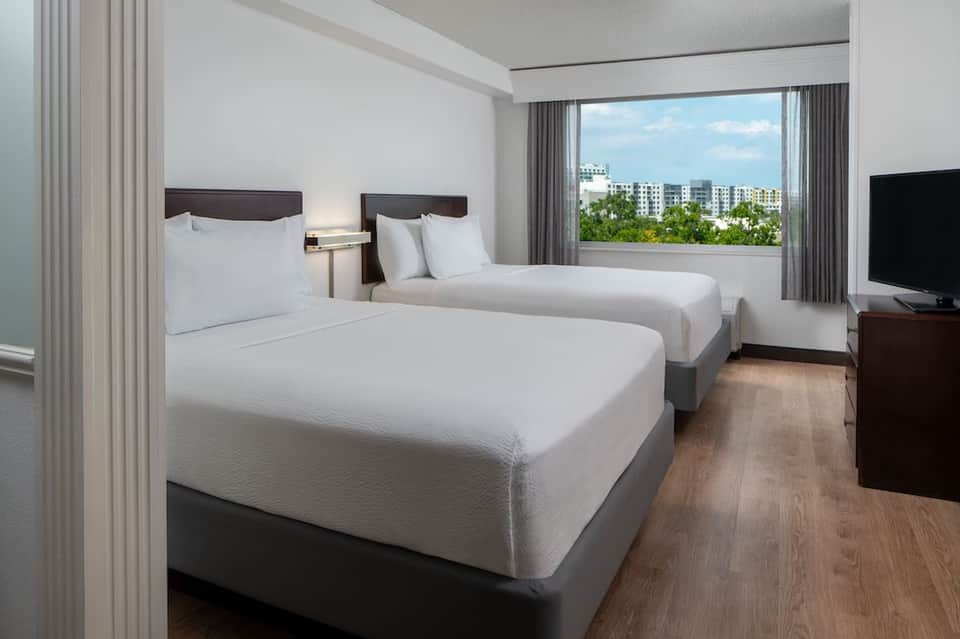 Modern hotel room with two queen beds, windows overlooking city skyline and residential buildings
