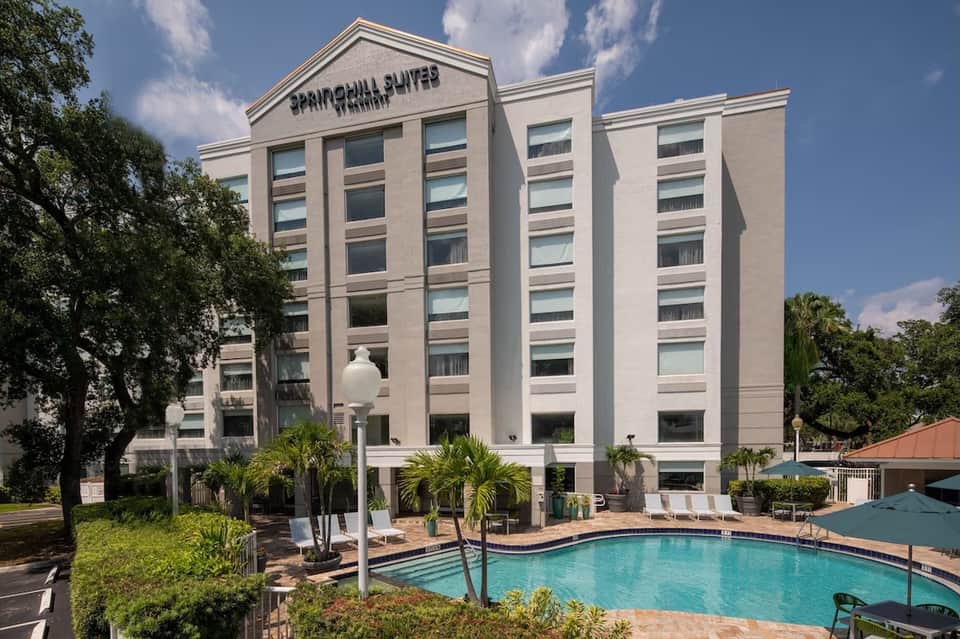 Beige multi-story hotel building with columns, pool in foreground, palm trees, and blue sky