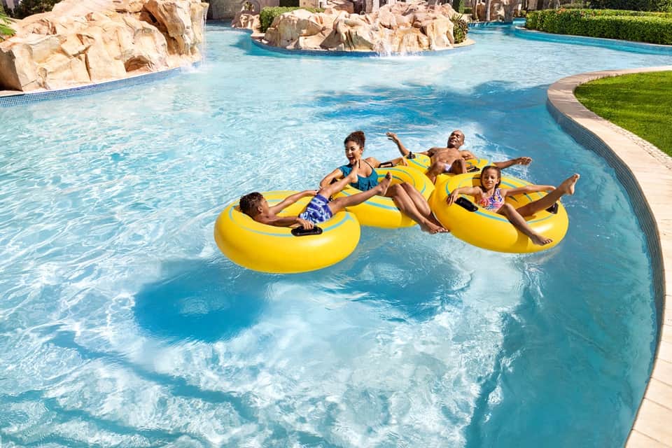Resort lazy river with families floating on yellow inflatables, rock formations, and lush green landscaping