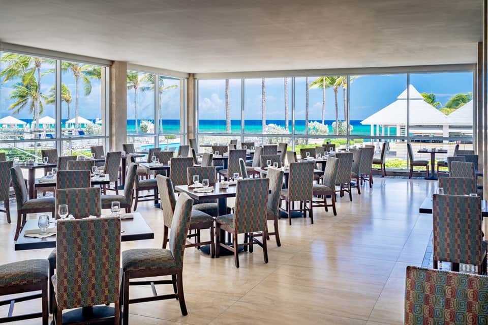 Fine dining restaurant with ocean and palm tree views through floor-to-ceiling windows, formal table settings