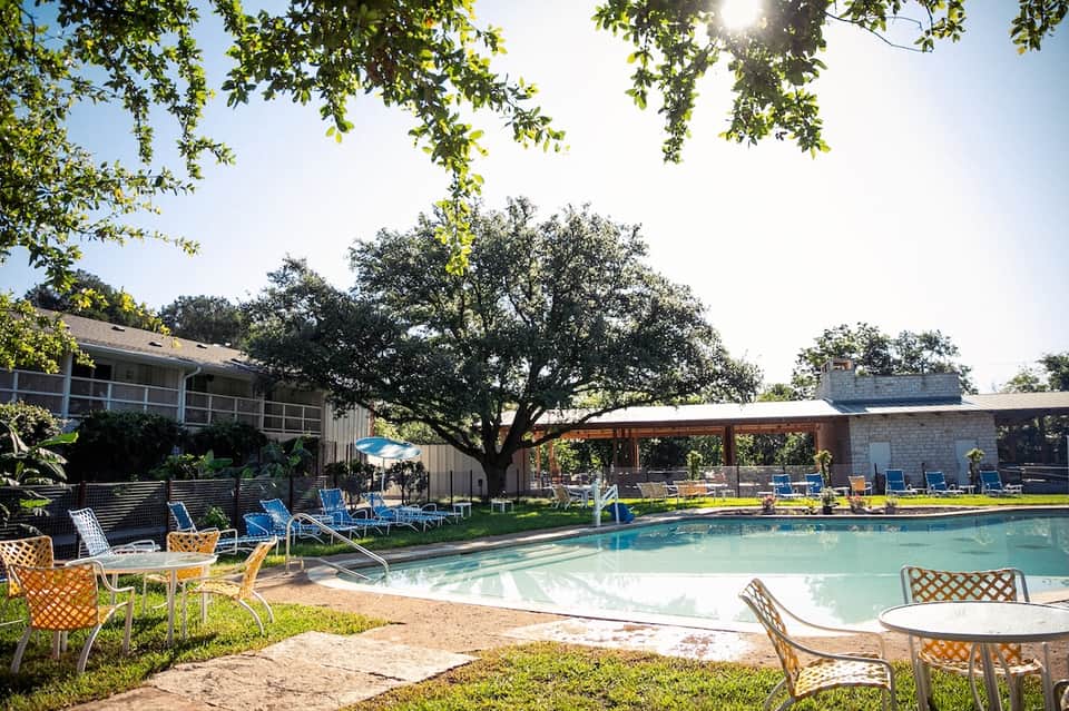 Resort pool area with loungers, shade structures, mature oak trees, and stone buildings on sunny day