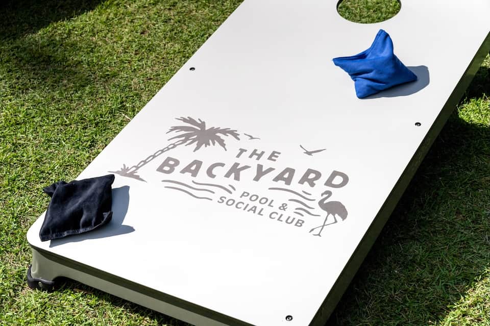 Cornhole game set on grass with 'The Backyard Pool & Social Club' logo