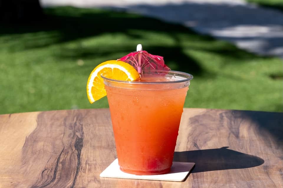 Tropical cocktail with orange slice and pink umbrella on wooden table by grass