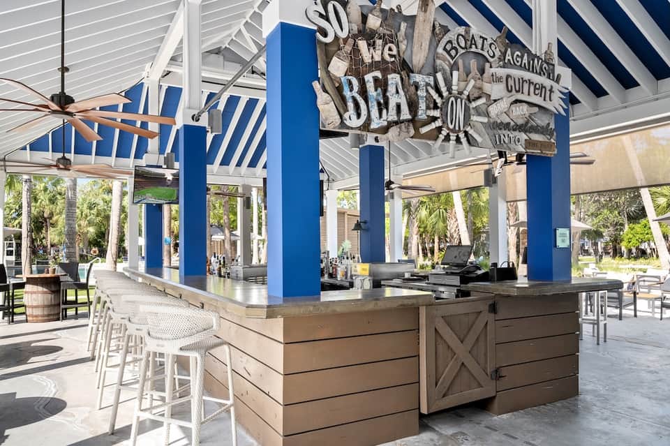 Beachfront tiki bar with blue columns, striped awning, white bar seating, nautical decor, and tropical surroundings