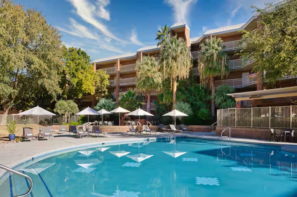 Resort pool with white umbrellas, lounge chairs, and multi-story brick hotel building