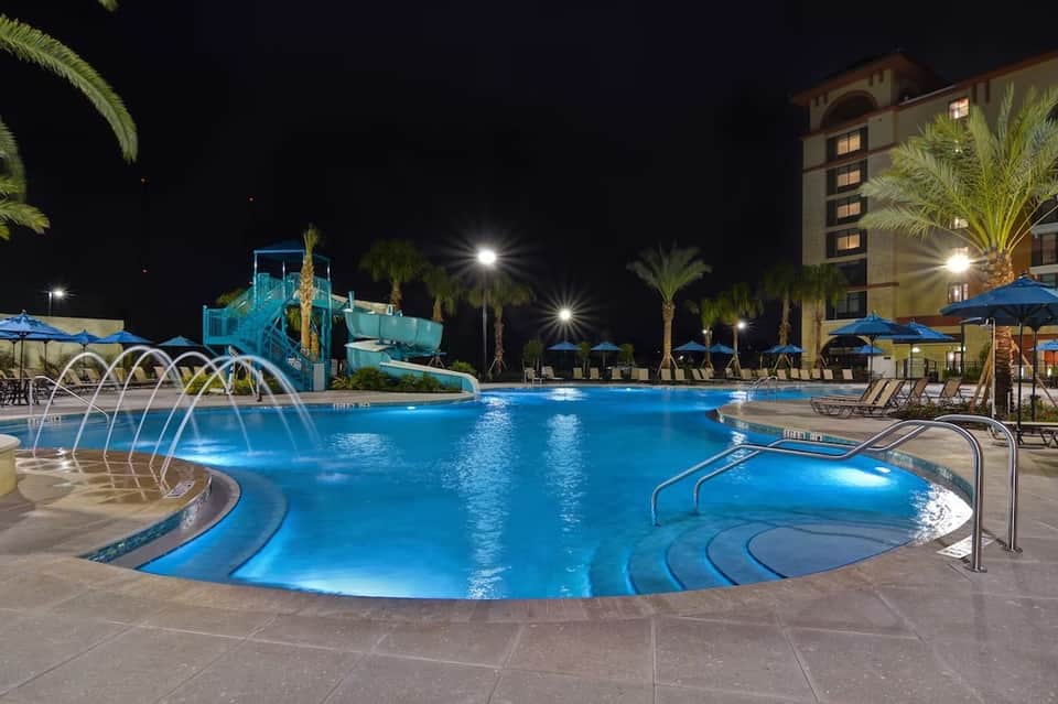 Illuminated resort pool at night with water slides, palm trees, and hotel building