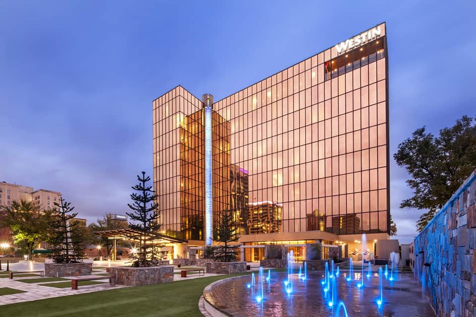 Modern hotel building exterior at dusk with illuminated glass facade and blue fountain in plaza