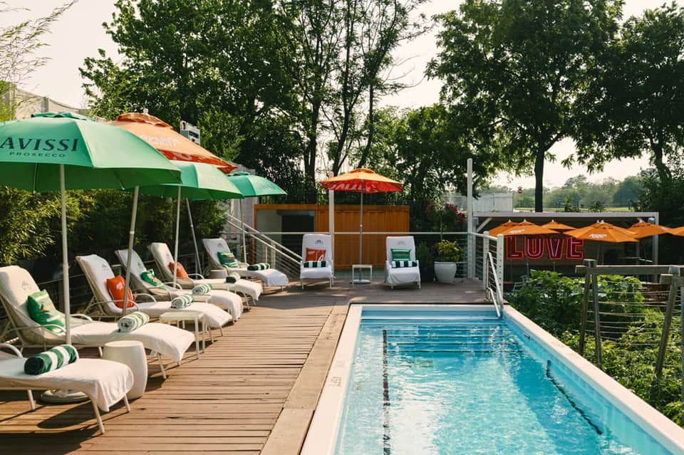 Outdoor deck with wooden loungers under green and orange umbrellas beside small swimming pool surrounded by trees