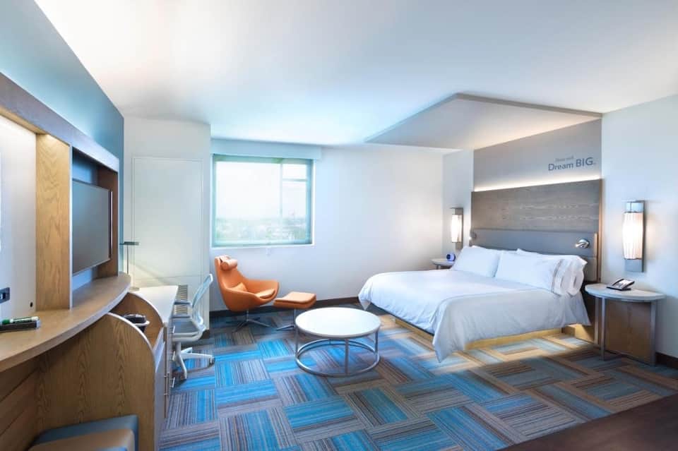 Spacious hotel suite with bed, lounge area, desk, orange chair, and floor-to-ceiling windows