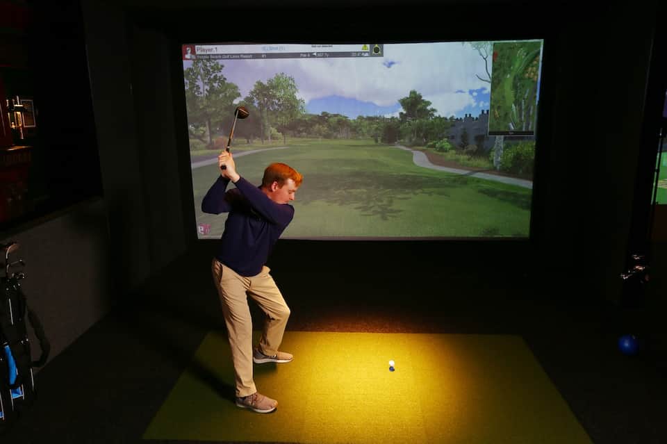 Man using golf simulator with large screen display showing virtual golf course gameplay