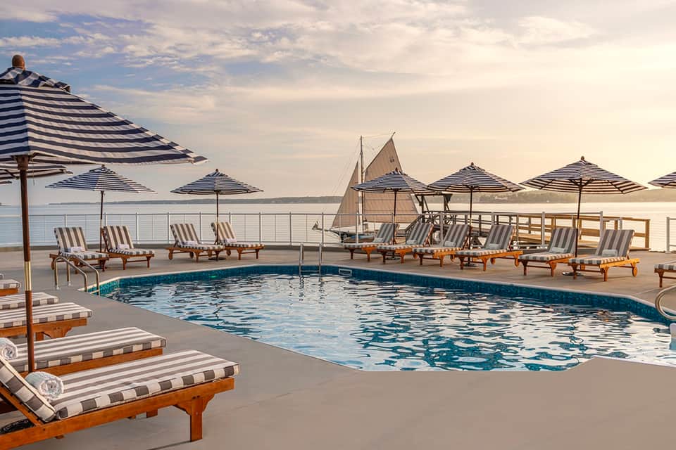 Oceanfront pool with sailboat decoration, striped umbrellas, lounge chairs, and horizon water views