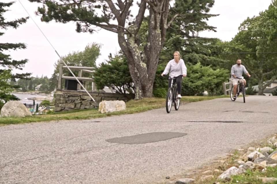 Two people cycling on a residential street near trees and stone structures in a quiet neighborhood
