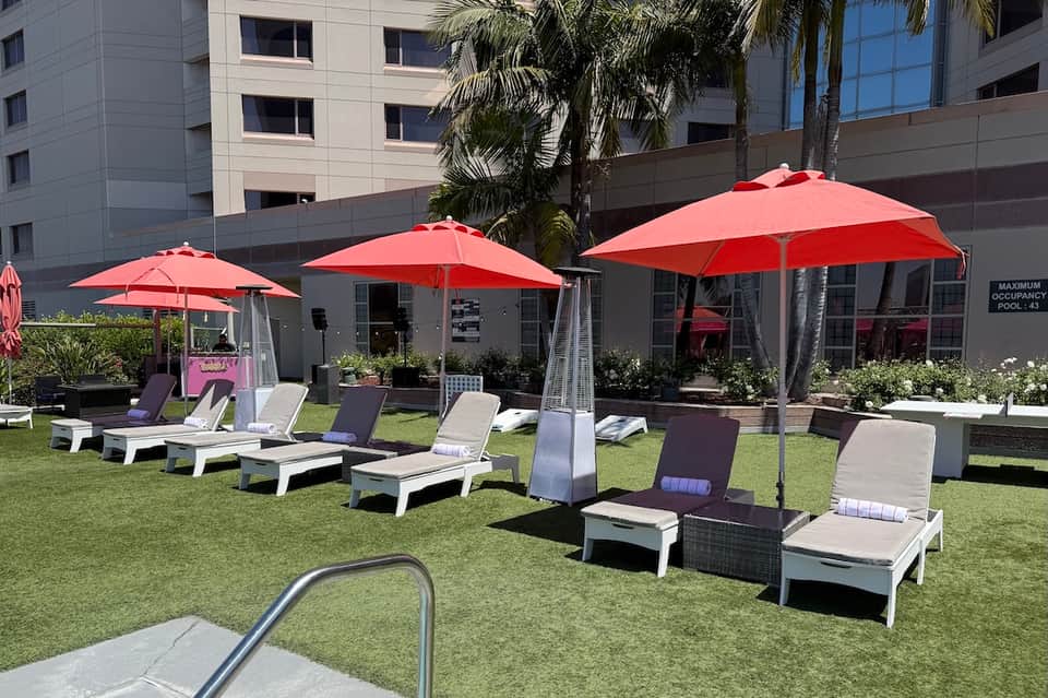 Resort lawn with white lounge chairs under red umbrellas, modern building, palm trees, and green grass