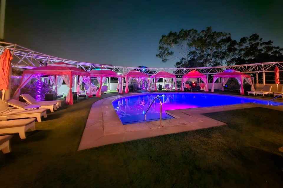 Nighttime pool area with pink and purple lighting, white cabanas, and lounge chairs