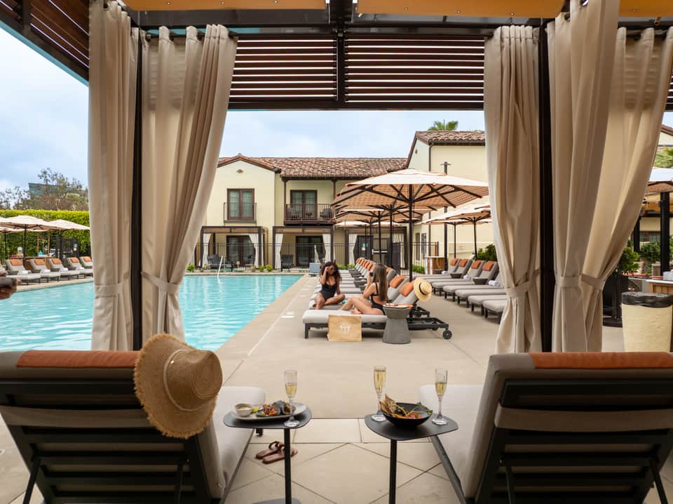 Luxury pool pavilion with cream curtains, lounge chairs, white umbrellas, and Spanish-style architecture visible