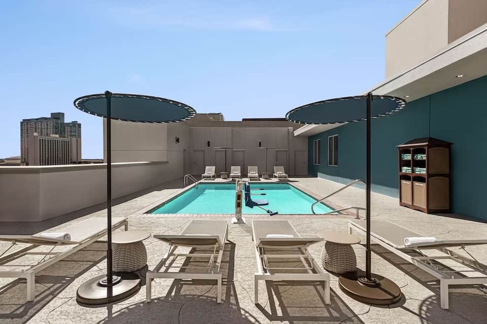 Rooftop pool area with turquoise lap pool, lounge chairs, umbrellas, and city skyline view