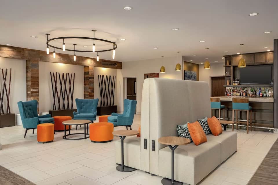 Modern hotel lobby lounge with turquoise chairs, orange ottomans, bar area, and pendant lighting