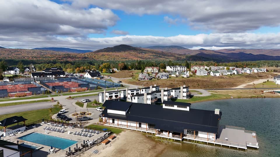 Aerial view of lakeside resort with pool, white hotel building, mountains, forests, and sports courts