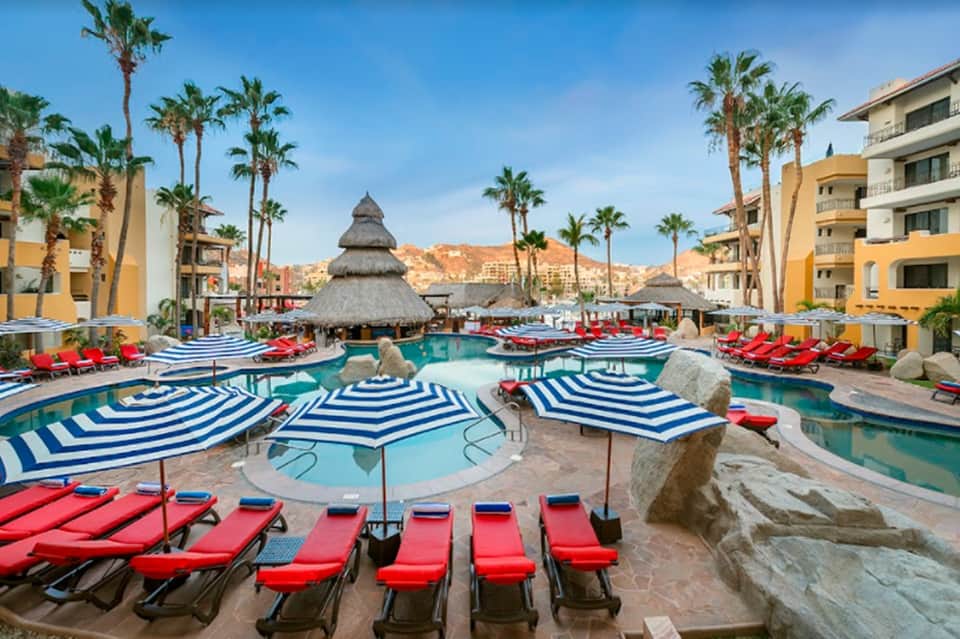 Resort pool area with blue and white striped umbrellas, red loungers, thatched roof structures, and surrounding buildings