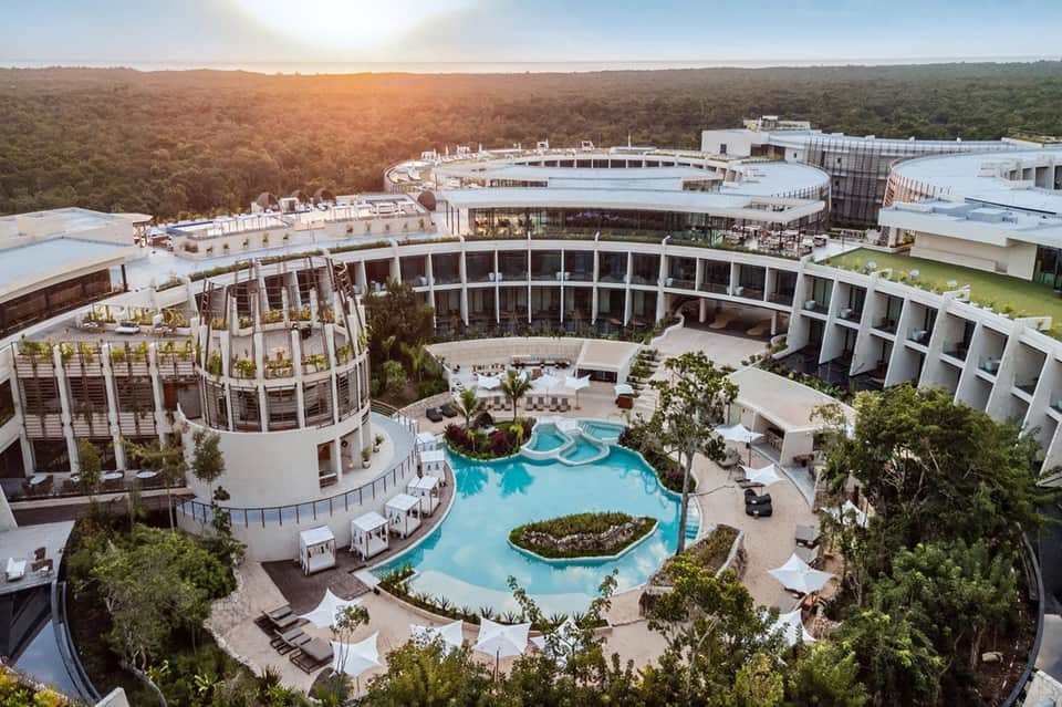 Aerial view of large resort with curved pool, modern architecture, and surrounding forest landscape