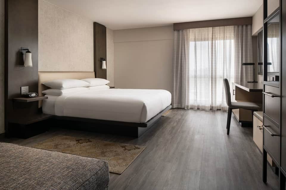 Modern hotel bedroom with white bedding, gray wood flooring, desk, and balcony with sheer curtains