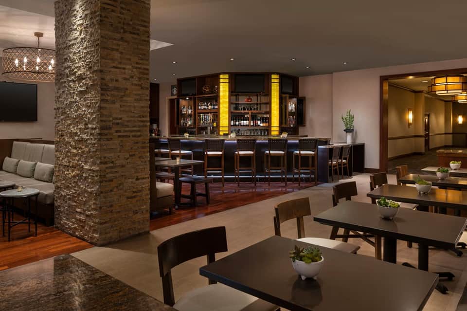 Upscale resort bar and lounge area with brick column, dark wood furnishings, and illuminated bar counter