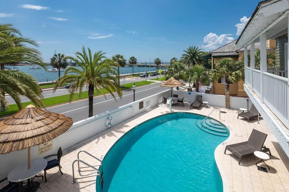 Curved swimming pool on waterfront patio with palm trees, ocean view, and white buildings