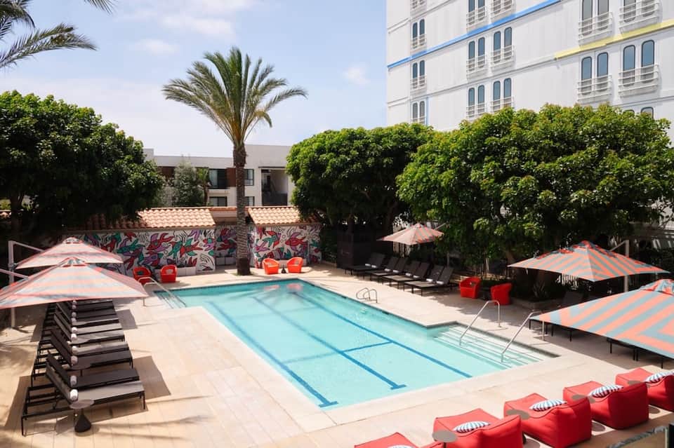 Resort pool with striped umbrellas, red seating, palm trees, and modern building backdrop