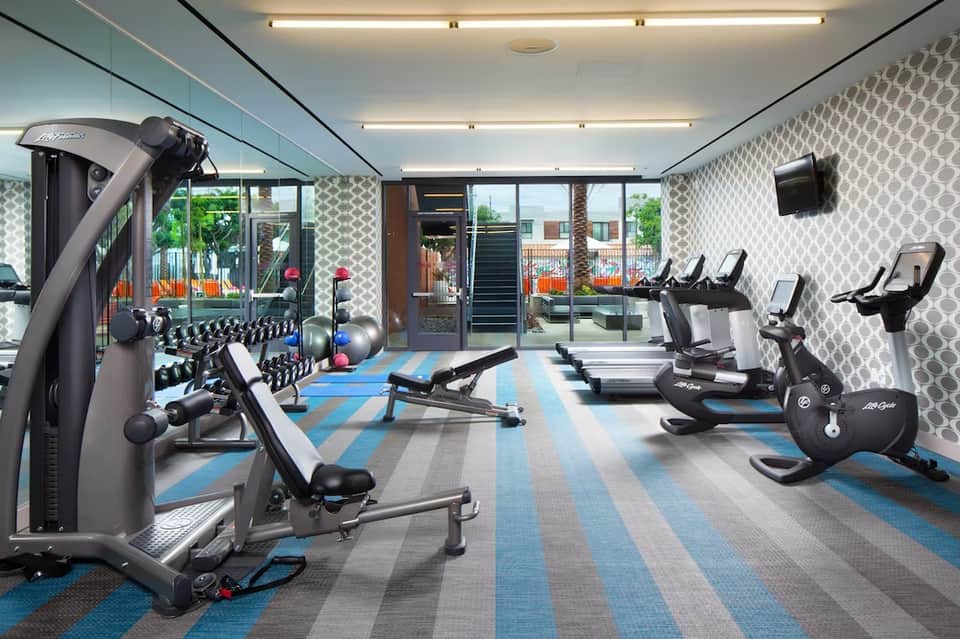 Fitness center with cardio equipment, free weights, blue striped flooring, and windows overlooking grounds