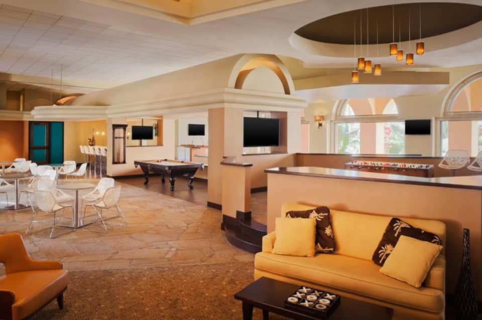 Luxurious hotel lounge with orange and cream décor, billiards table, comfortable seating, and multiple flat-screen TVs