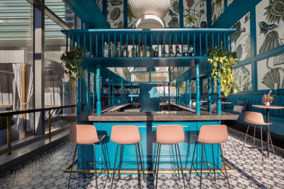 Modern bar interior with teal blue columns, botanical wallpaper, and coral seating