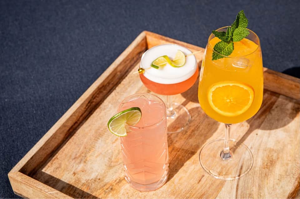 Three colorful cocktails with citrus garnishes on wooden serving tray against dark background
