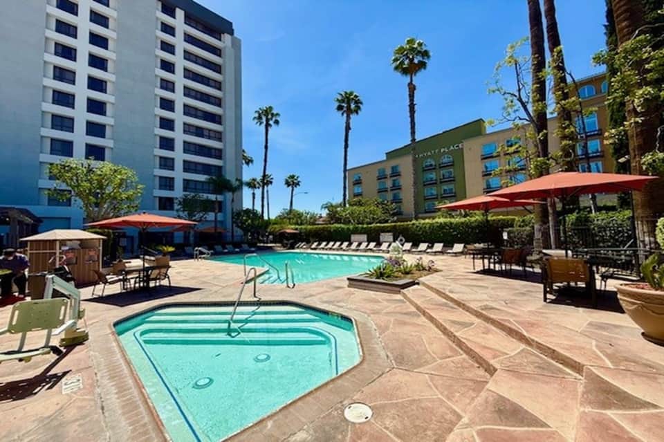 Resort pool area with sun loungers, umbrellas, and multi-story hotel buildings with palm trees