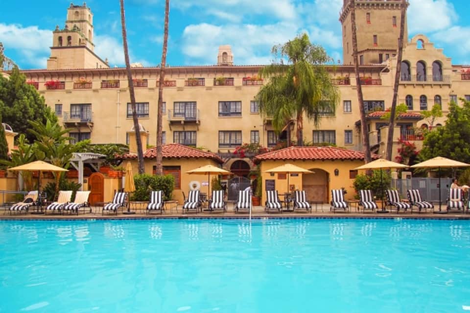 Historic Spanish-style hotel with yellow stucco facade overlooking large swimming pool with lounge chairs and umbrellas
