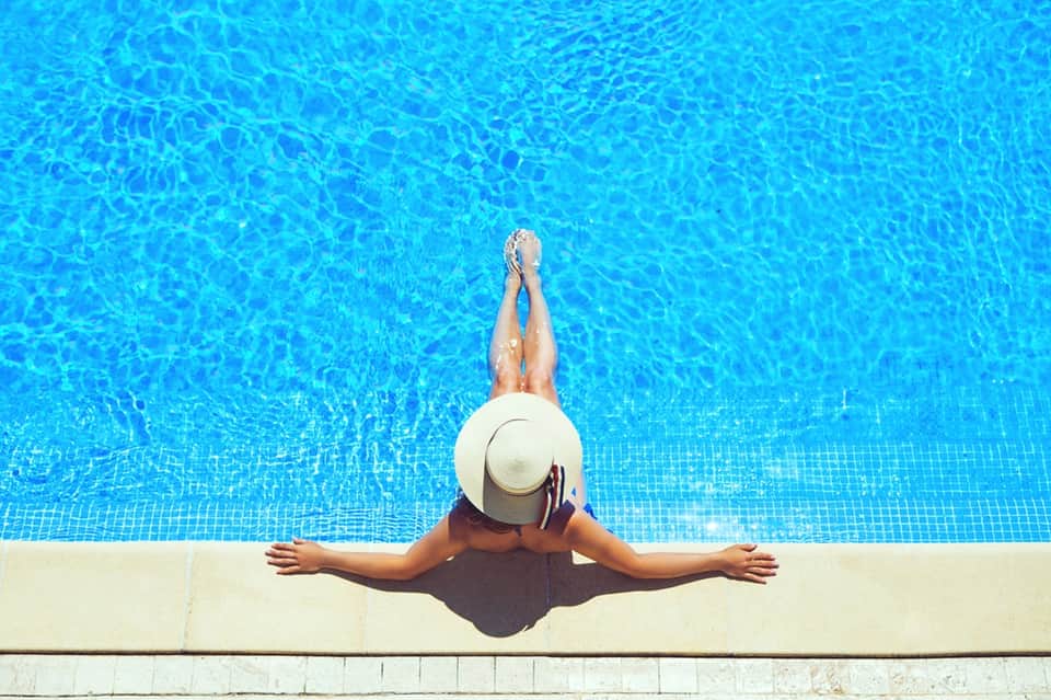 Woman in white hat sitting at pool edge with legs in bright blue water