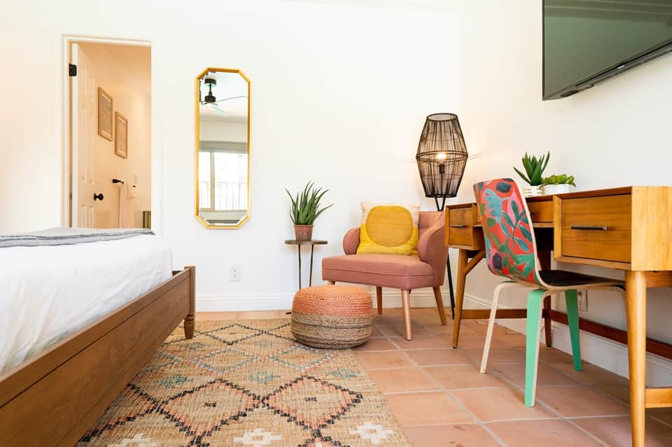 Mid-century modern hotel room with wooden bed, colorful accent chair, plants, and rustic decor