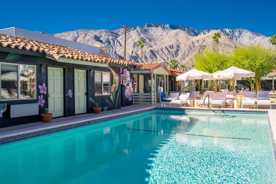 Desert resort pool with turquoise water, white umbrellas, mountain backdrop, and colorful floral painted buildings