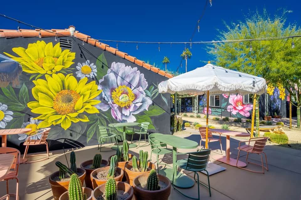 Desert spa courtyard with colorful flower mural, white umbrella, potted cacti, and vibrant outdoor furniture