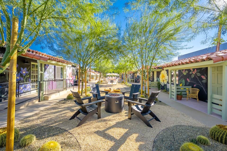 Desert resort courtyard with colorful bungalows, fire pit, mature trees, and blue sky