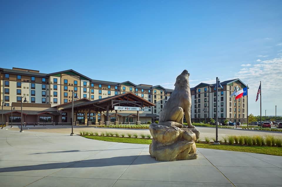 Great Wolf Lodge exterior with tall bear statue, building entrance, flags, and manicured grounds on sunny day