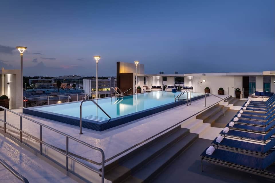 Modern rooftop pool at dusk with lounge chairs, glass railings, and city skyline view