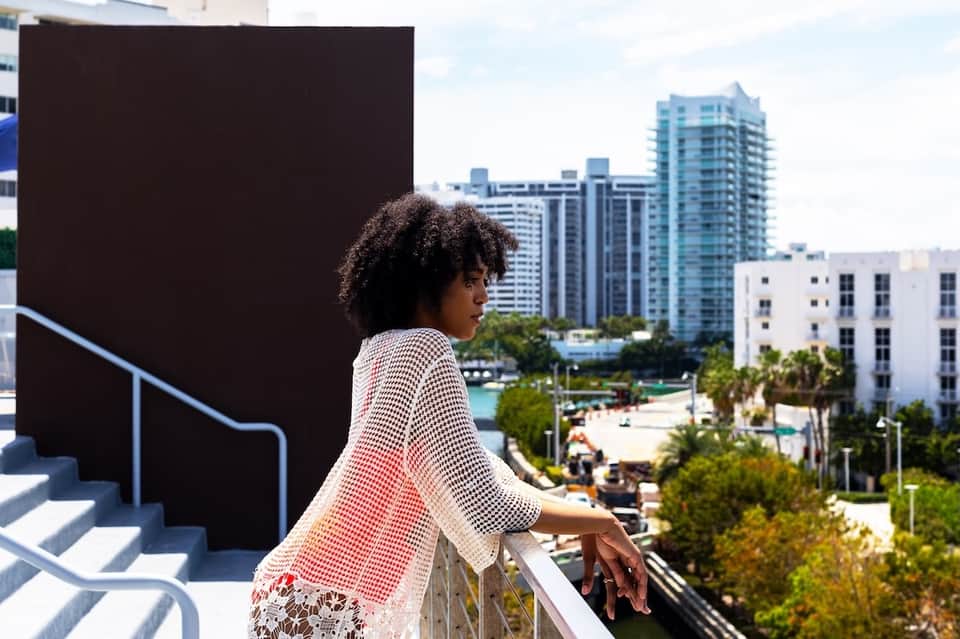 Woman in patterned jacket on hotel balcony overlooking waterfront and modern cityscape