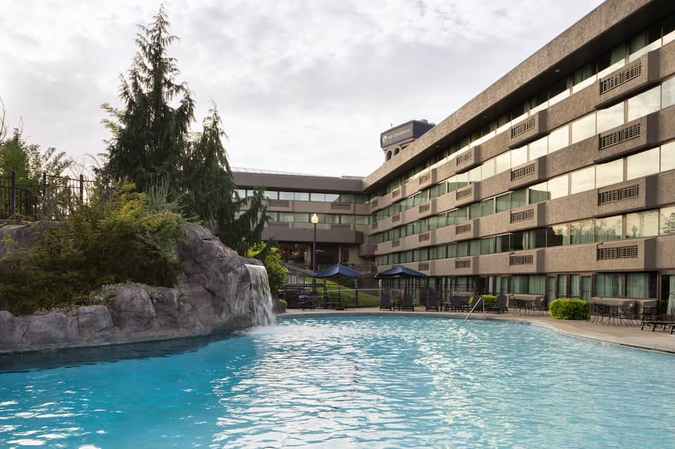 Outdoor heated swimming pool with natural waterfall feature, hotel building and evergreen trees in background